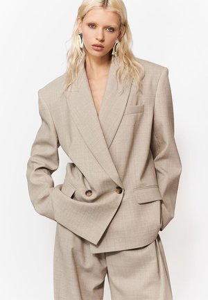 Blondynka o falowanych włosach, ubrana w beżową, dużą dwurzędową marynarkę i pasujące szerokie spodnie, stojąca z rękami w kieszeniach.