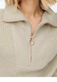 Pull en maille beige avec un col zippé à mi-hauteur, présentant un motif texturé et un curseur de fermeture éclair en forme d'anneau doré.