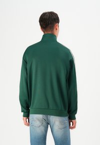 Veste zippée verte avec un col haut, des rayures latérales blanches et des poignets côtelés, associée à un jean en denim bleu clair et délavé. Texture de tissu lisse.