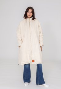 Długa, kremowa kurtka z zamkiem z przodu, kapturem i bocznymi kieszeniami. Na dole ma pomarańczową metkę. Nosi się ją na rozkloszowanych dżinsach i białych butach.