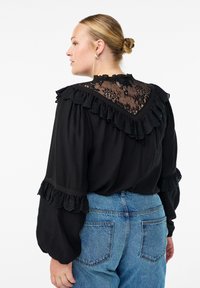 Frau mit blondem Dutt, die eine schwarze Bluse mit Spitze und Rüschen sowie hochgeschnittene blaue Jeans trägt und sich leicht zur Seite dreht.