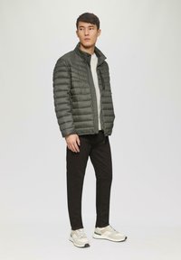 Padded olivgrüne Jacke mit Reißverschluss, kombiniert mit einem hellgrauen Oberteil, schwarzen Hose und beigen Turnschuhen mit grünen Akzenten.