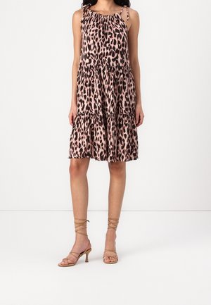 Femme portant une robe rose à imprimé léopard sans manches, longueur genou, avec une jupe à volants, et des sandales à talons à brides couleur caramel, sur fond blanc.