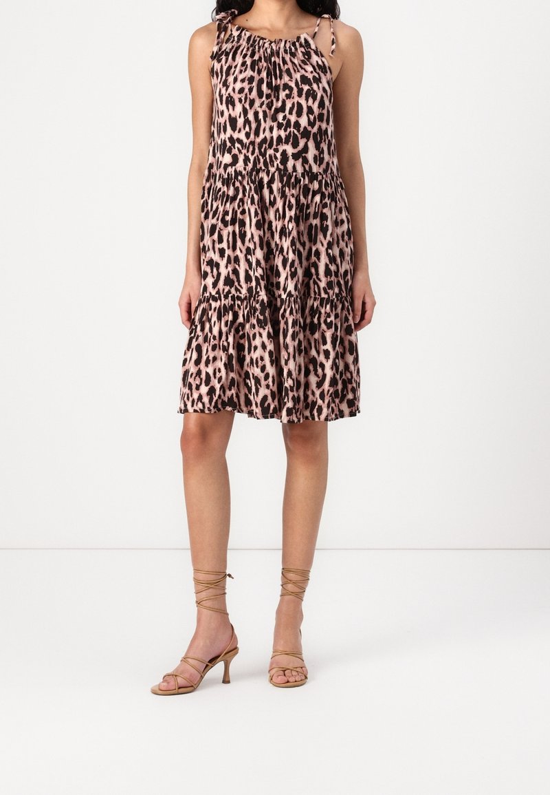 Femme portant une robe rose à imprimé léopard sans manches, longueur genou, avec une jupe à volants, et des sandales à talons à brides couleur caramel, sur fond blanc.