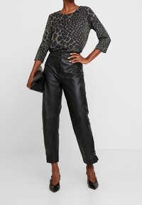 Zwarte leren broeken met een getailleerde vorm, gecombineerd met een groen en zwart dierenprint blouse. Het model houdt een zwarte clutch vast.