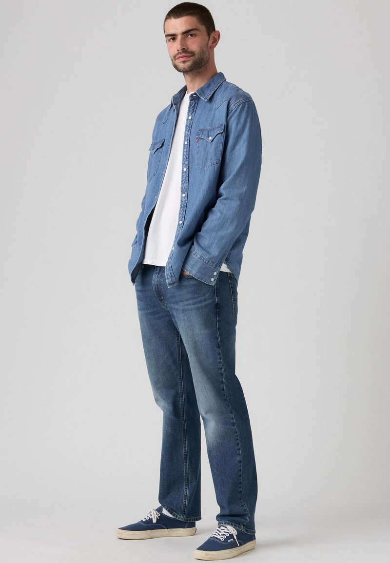 Man standing with hands in pockets wearing blue denim shirt, white t-shirt, blue jeans, and blue sneakers against a plain light background.