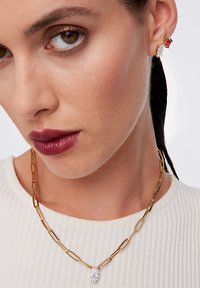 Złoty naszyjnik łańcuszkowy z wydłużonymi ogniwami oraz fasetowanym przezroczystym wisiorkiem. Modelka ma na sobie małe kolczyki wkręty z czerwonymi i przezroczystymi kamieniami szlachetnymi.