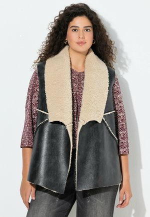 Gilet di pelle nera con colletto in shearling beige, davanti aperto e cuciture in crema a contrasto. Indossato sopra un top a motivo borgogna.