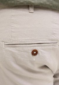 Poche de pantalon beige texturée avec un seul bouton marron et une boucle de ceinture visible en haut.