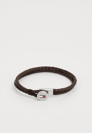 Bracelet en cuir tressé marron avec un fermoir en argent arborant un logo circulaire, agrémenté d'éléments rouges et bleus.