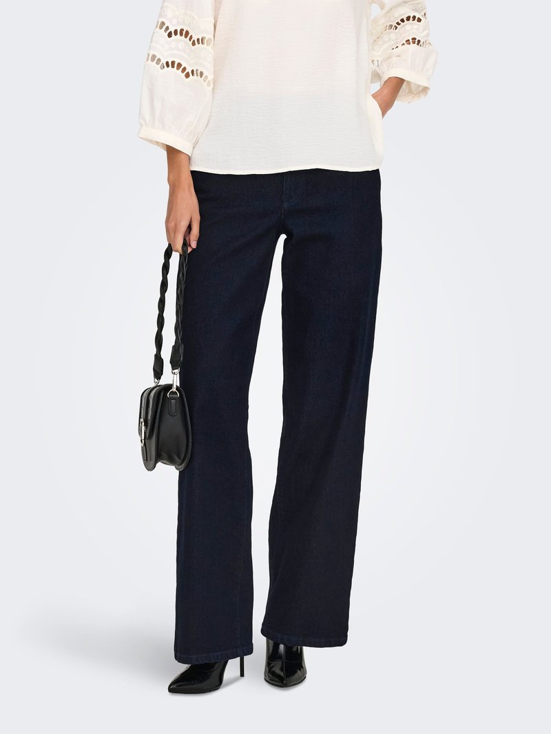 Femme portant un jean large foncé, un chemisier blanc avec des détails en broderie anglaise, des bottes noires à talons hauts, et tenant un petit sac à main noir.