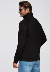 Chaqueta negra con cremallera y cuello alto, de tejido texturizado, mangas largas y puños acanalados. Lleva puesta con vaqueros de color azul claro y gafas de sol.