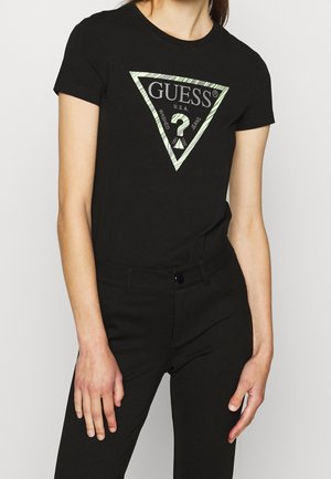 Persona con una camiseta negra de Guess con un logo triangular y pantalones negros, de pie con los brazos relajados frente a un fondo claro.