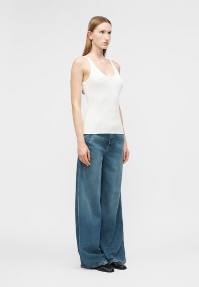 Γυναίκα που φοράει λευκό αμάνικο τοπ με V-λαιμόκοψη, φαρδιά μπλε τζιν και μαύρα μυτερά παπούτσια, στέκεται μπροστά σε απλό φόντο.