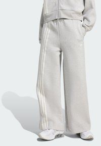 Pantalones de chándal grises hechos de una suave tela, con piernas anchas y rayas verticales blancas a los lados. Llevados con zapatillas blancas.