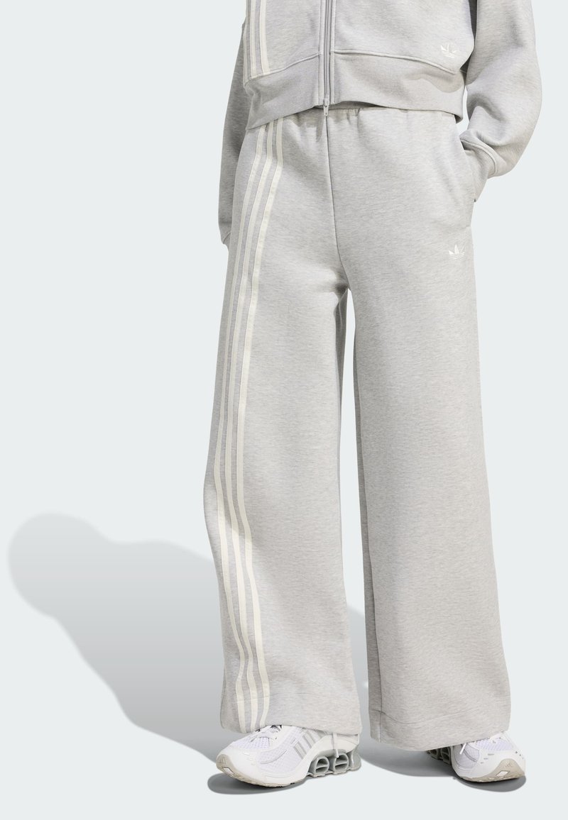 Pantalones de chándal grises hechos de una suave tela, con piernas anchas y rayas verticales blancas a los lados. Llevados con zapatillas blancas.