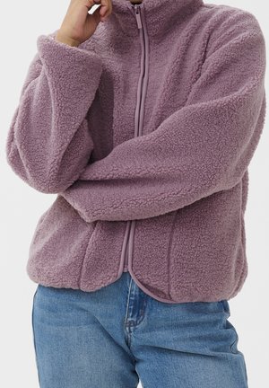 Person iført lys lilla, fluffy lynlåstrøje og blå jeans, med den ene arm krydset og hånden hvilende ved hagen.
