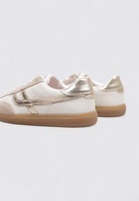 Zapatillas blancas y doradas con una parte superior de ante y cuero, diseñadas con un acabado suave, cordones planos y una suela de goma color chicle.