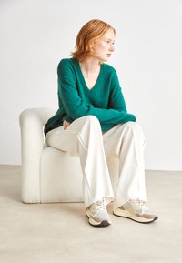 Grüner, V-Ausschnitt, flauschiger Pullover, kombiniert mit lockeren, cremefarbenen Hosen und beigen Sneakers. Das Outfit ist lässig gestylt, während es auf einem weichen, runden Stuhl sitzt.