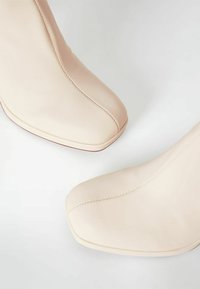 Botas altas de tacón en tono nude, hechas de cuero liso, con punta cuadrada, detalles de costuras visibles y un diseño contorneado elegante.