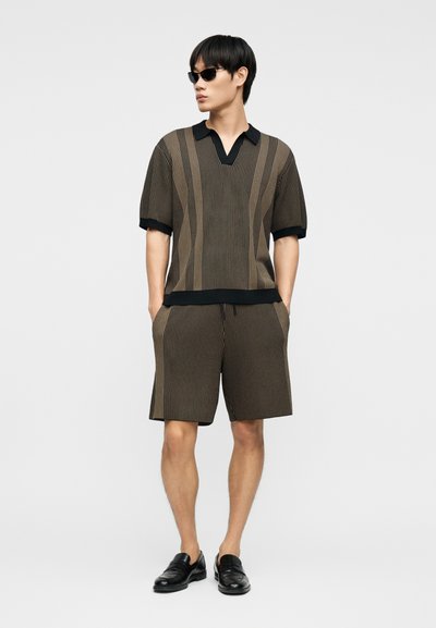 Homme portant des lunettes de soleil noires et une chemise à manches courtes marron à fines rayures, assortie à un short, avec des mocassins noirs, les mains dans les poches.