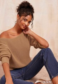 Hellbrauner Strickpullover mit schulterfreiem Design, kombiniert mit blauen Jeans. Das Model hat lockige Haare und trägt goldene Creolen.