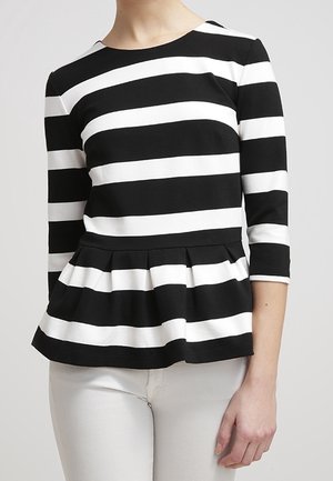 Kobieta ubrana w czarno-biały, poziomo paskowany top peplum z rękawami do łokcia i białe spodnie, stojąca na jednolitym tle.
