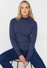 Navyfarbener, gerippter Rollkragenpullover mit langen Ärmeln, der über Knopfakzente an der Schulter und am Bündchen verfügt. Kombiniert mit dunkelblauen Jeans.