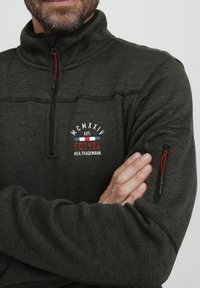Pull en polaire vert foncé avec un col haut, fermeture éclair half-zip et tissu texturé. Doté d'accents rouges et d'un logo brodé sur la poitrine.