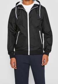 Zwarte windjack met een rits aan de voorkant, witte accenten, een verstelbare capuchon en elastische manchetten. Gecombineerd met marineblauwe broek.