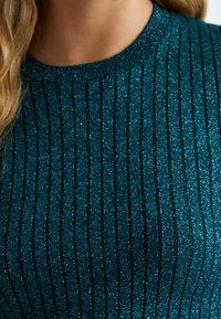 Türkisfarbenes, geripptes Strickoberteil mit glänzenden Metallicfäden. Besitzt einen runden Ausschnitt und auffällige vertikale Streifen. Weiche Textur.
