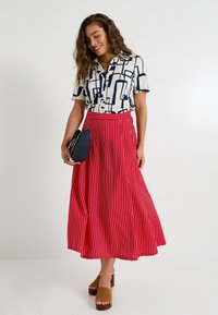 Chemise blanche avec un motif géométrique bleu marine, jupe maxi rayée rouge et blanche, sandales à talons épais marron, et sac à bandoulière noir.
