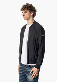 Giubbotto bomber nero realizzato in tessuto leggero con polsini e colletto a coste, dotato di una cerniera frontale e di un piccolo emblema sulla manica.