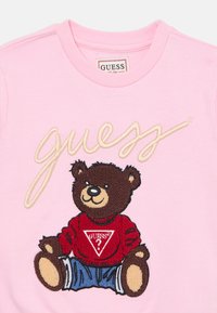 Felpa rosa con un orsetto ricamato che indossa un maglione rosso con un logo triangolare e pantaloni corti blu. Il logo "Guess" è ricamato sopra.