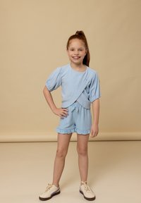 Lichtblauw tweedelig outfit met pofmouwen, gerimpelde short en een wikkelshirt. Draagt beige sneakers op een neutrale achtergrond.