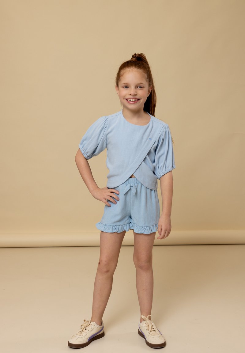 Lichtblauw tweedelig outfit met pofmouwen, gerimpelde short en een wikkelshirt. Draagt beige sneakers op een neutrale achtergrond.