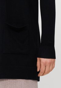 Zwarte gebreide cardigan met geribde manchetten en zoom. Bevat een voorzak met een gestructureerde afwerking, weergegeven met een hand die ernaast rust.