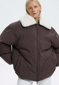 Puchowa kurtka w ciemnym brązie z białym sztucznym futrem przy kołnierzu, z zamkiem błyskawicznym i bocznymi kieszeniami. Gruby materiał o fakturowanej powierzchni.