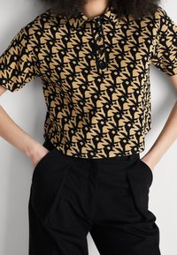 Polo à manches courtes beige et noir à motifs avec boutons, porté rentré dans un pantalon noir taille haute, mains dans les poches.