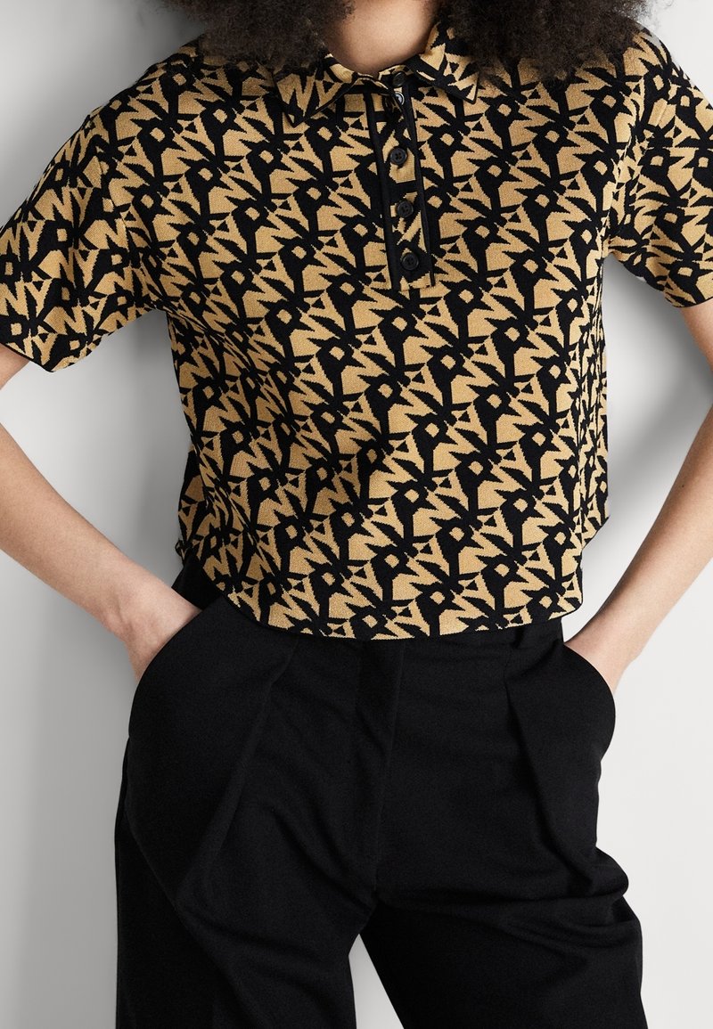 Polo à manches courtes beige et noir à motifs avec boutons, porté rentré dans un pantalon noir taille haute, mains dans les poches.