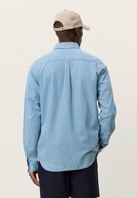 Ανοιχτό μπλε πουκάμισο από τζιν με μακριά μανίκια, πατιλέτα με κουμπιά και καμπυλωτό τελείωμα. Διαθέτει λοξό πλάτη και μανσέτες με κουμπιά.