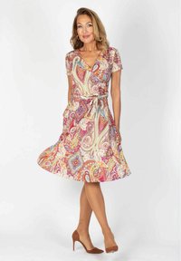 Kvinde iført en knælang, kortærmet kjole med farverige paisleymønstre, brune høje hæle og guldringe, stående foran en hvid baggrund.