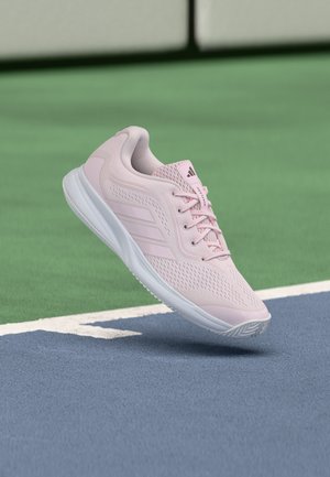 Chaussure de sport rose clair avec semelle blanche flottant au-dessus d’une surface de court de tennis bleue et verte.