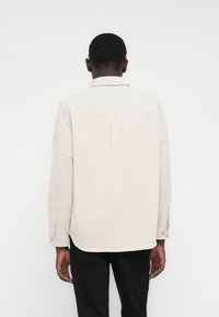 Világos bézs ing texturált anyaggal, gombos mandzsettával és hátul pliszírozott résszel. Gallérral és ívelt szegélyű kialakítással rendelkezik.