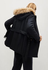 Parka noire avec capuche doublée de fourrure, taille ceinturée et poches latérales ; présente une texture lisse avec un design structuré.