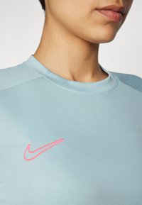 Vaaleansininen urheilupaita, jossa on pyöreä kaula-aukko ja vaaleanpunainen Nike-logo rinnassa. Kangas näyttää sileältä ja joustavalta.