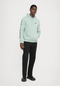 Light green hoodie made of soft fabric with a front pocket and drawstrings; features black logo and text on the left sleeve. Paired with black pants.