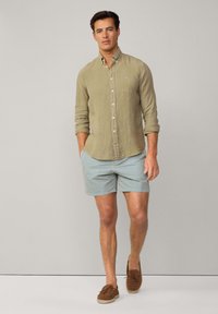 Leinenhemd mit Knöpfen in Olivgrün, kombiniert mit hellblauen gemusterten Shorts. Braune Slipper runden das Outfit ab. Einfaches, lässiges Design.