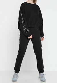 Schwarzer Sweatshirt mit weißem Grafikdesign an den Ärmeln, kombiniert mit schwarzen Hose. Das Material wirkt weich, mit einem lässigen Schnitt und elastischen Bündchen.