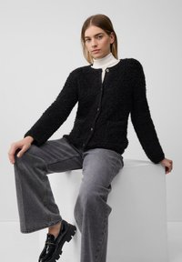 Schwarze strukturierte Strickjacke mit langenÄrmeln, rundem Ausschnitt, zwei Vordertaschen und Knopfleiste, kombiniert mit grauen weit geschnittenen Jeans und schwarzen Slippers.